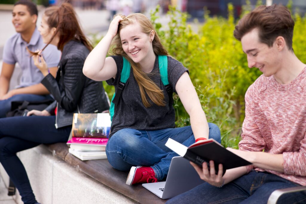 Female and male students outside of college, the female using her laptop and laughing and the boy reading a book