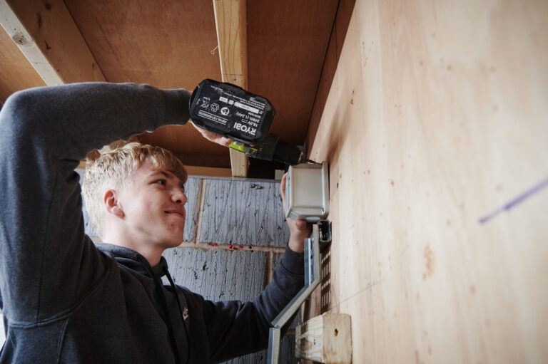 A male student smiling whilst using an electric screwdriver in a workshop at Herefordshire College