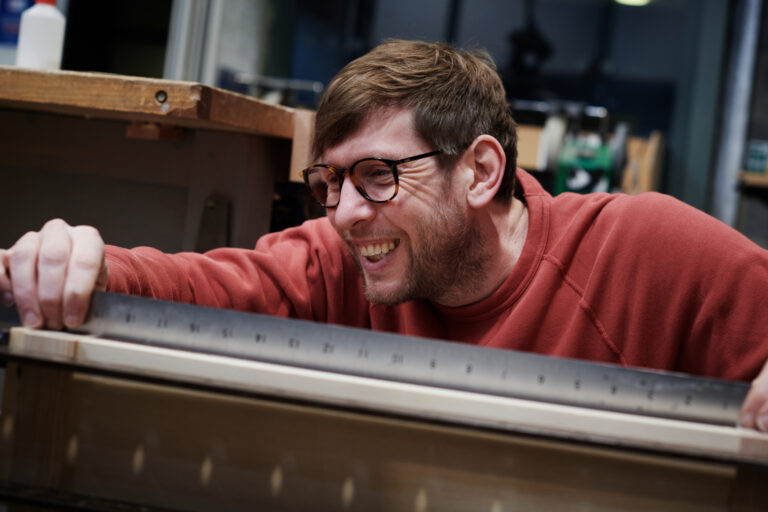 A male teacher smiling whilst demonstrates on measuring with woodwork at Herefordshire College