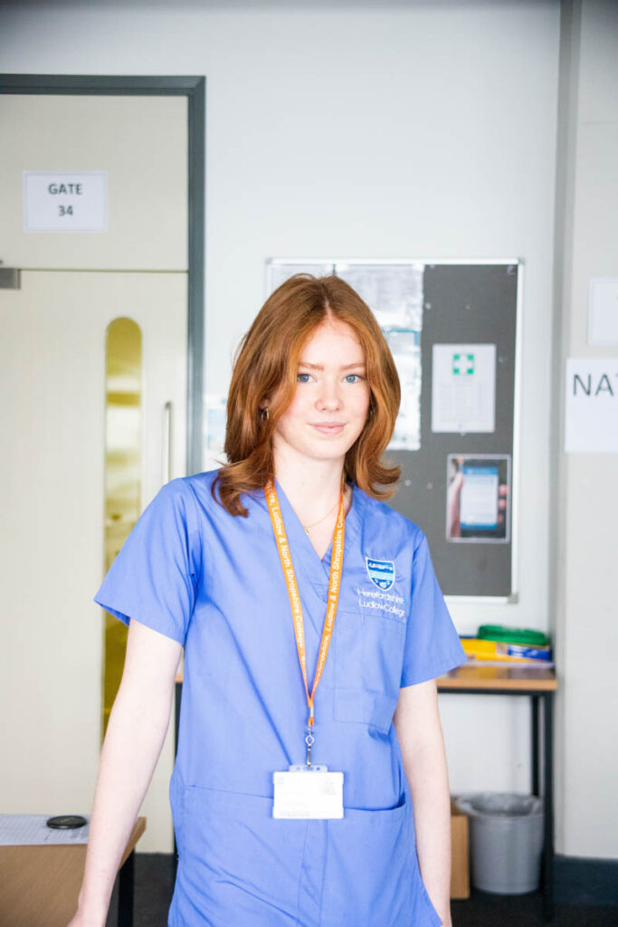 A student in HLNSC scrubs poses for a photo in a clinical setting within Herefordshire college.