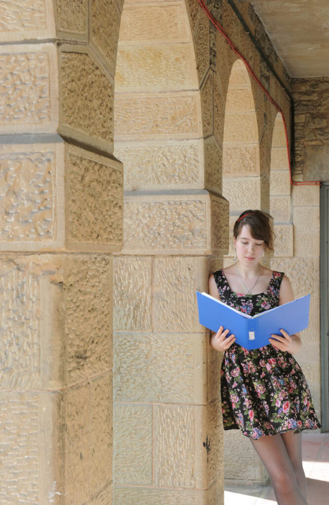 Girl leaning up against a sandstone wall reads from a blue folder.