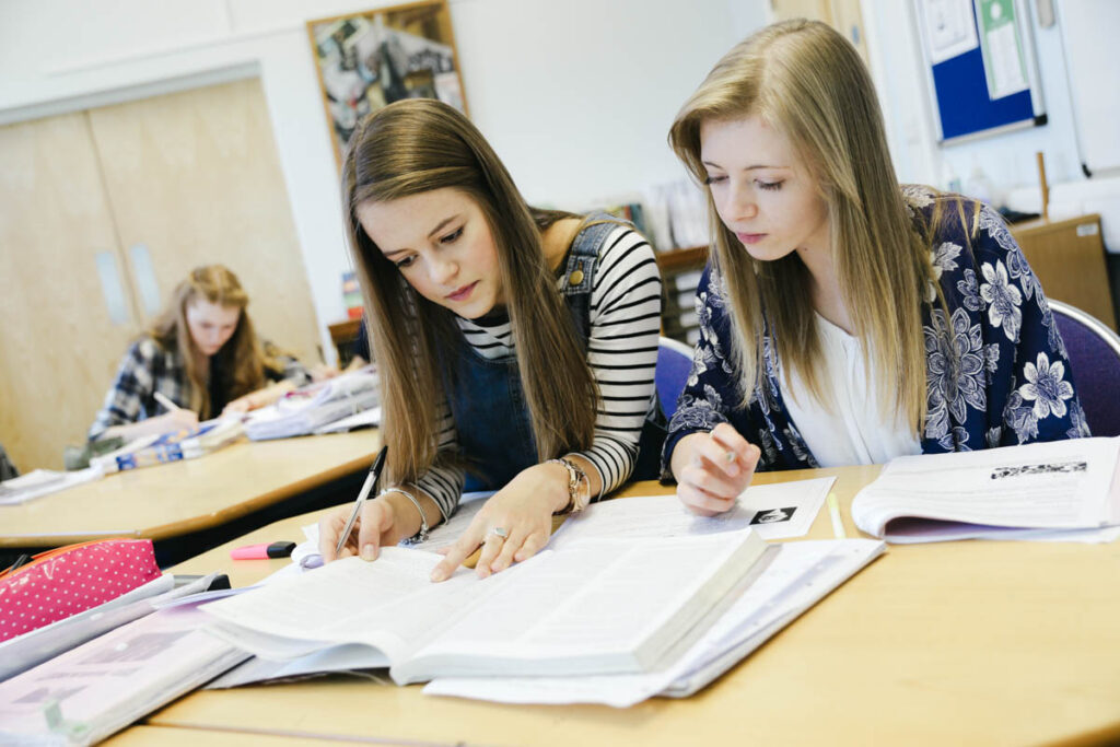 Two female students read from a text book in a classroom whilst completing a work book at the HLNSC Ludlow College.