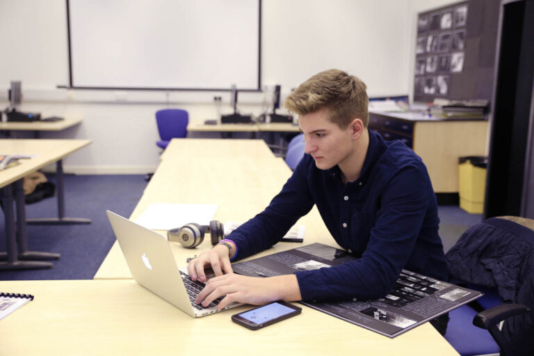 A student working in a portfolio book whilst using a macbook in a classroom at the HLNSC Ludlow College.