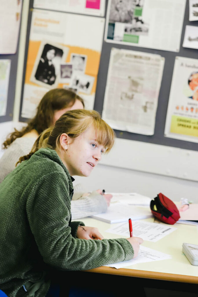 A female student writes on a sheet of paper whilst looking at something out of frame in a classroom at the HLNSC Ludlow College.