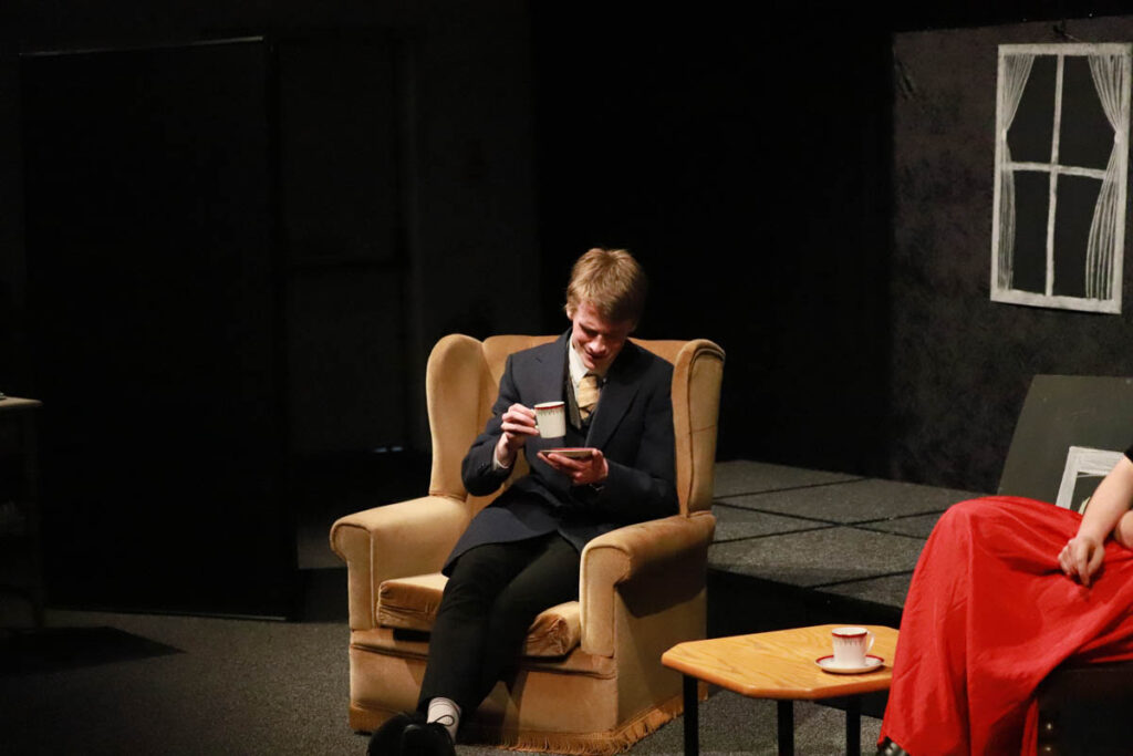 A male student is acting on stage dressed in costume sat in an arm chair at the HLNSC Ludlow College.