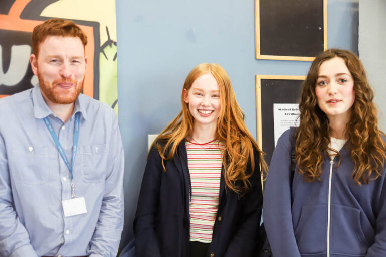 One male teacher smiling, two female students smiling in a classroom environment at Ludlow College