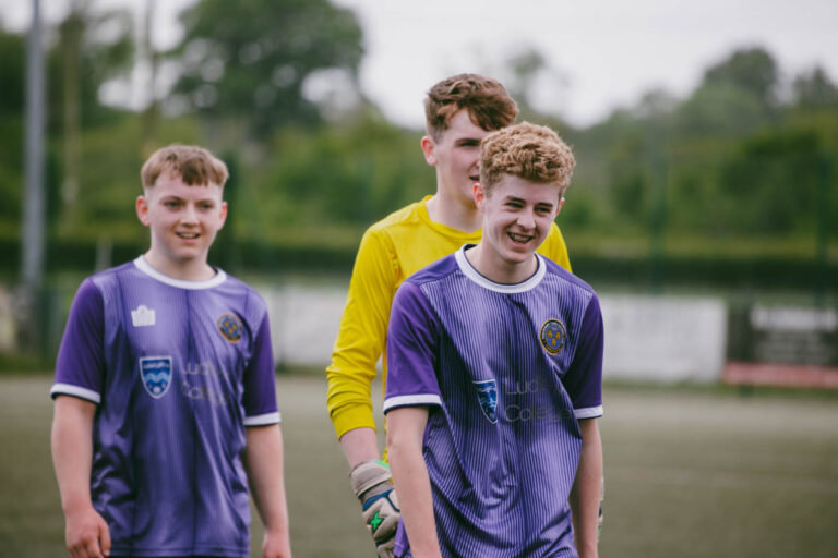 Three male students smiling on a pitch, two in purple kits and one in a yellow goalkeeper kit at Ludlow College