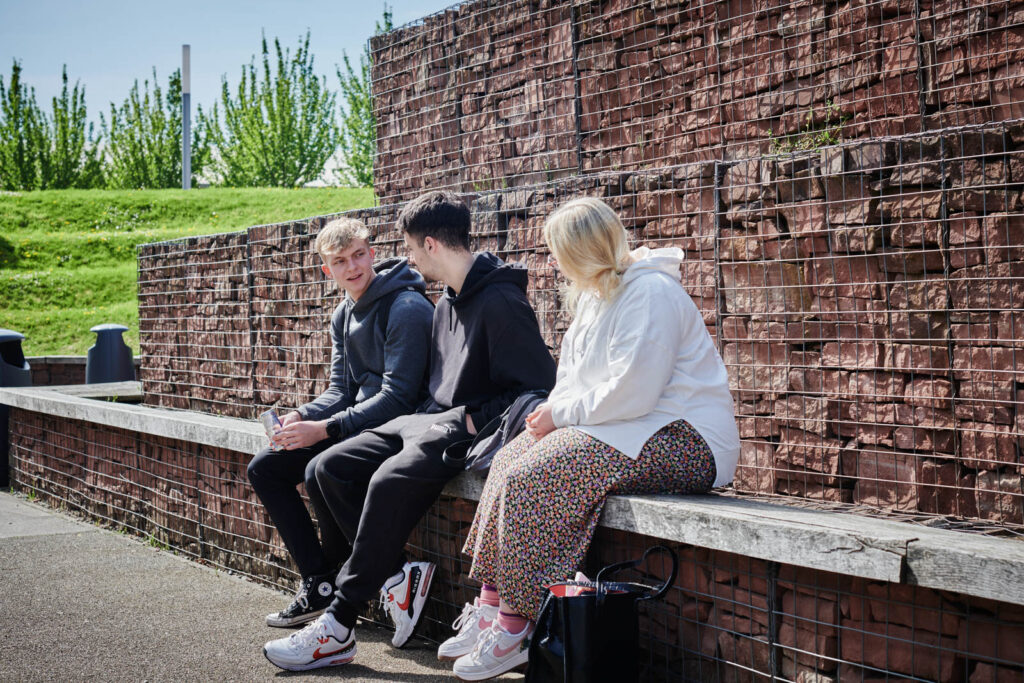 Two males and a female sat on a wall and talking outside Herefordshire college, enjoying the sunshine