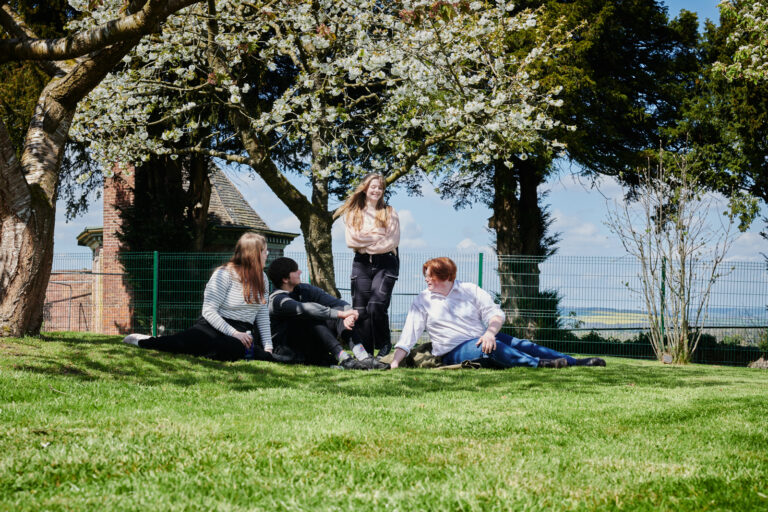 Three students sat and one stood laughing and talking on the grass in the sun outside Ludlow College