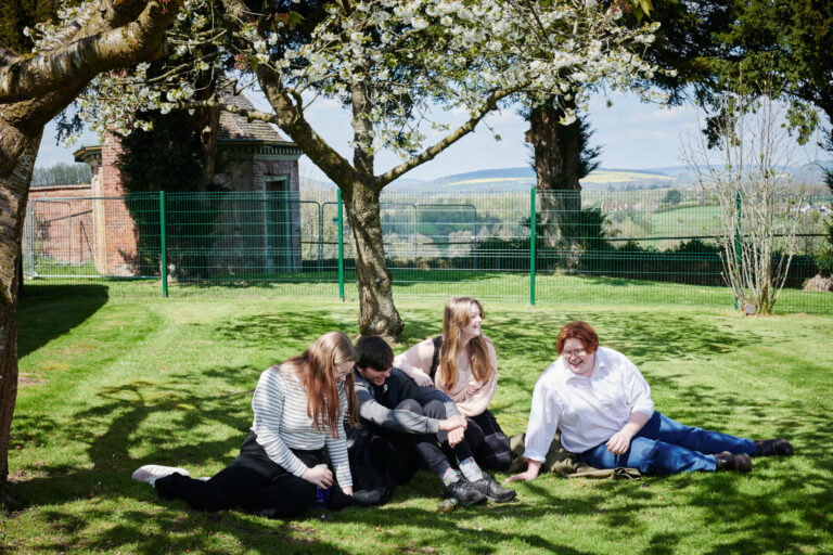 Four students sat laughing and talking on the grass in the sun outside Ludlow College
