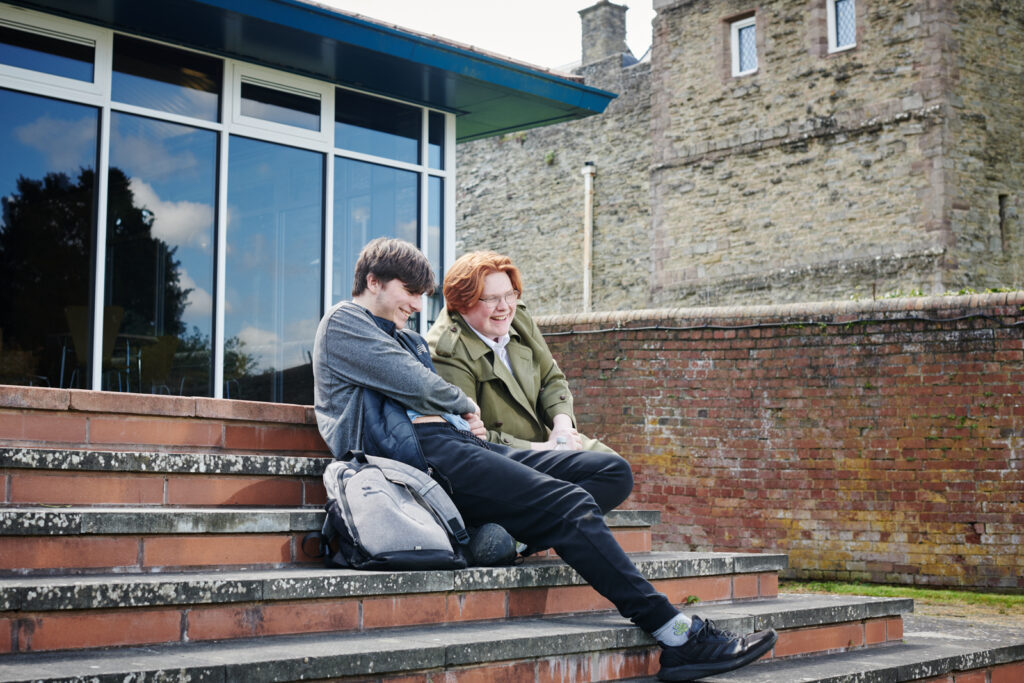 Two male students sat on stairs, laughing and talking outside Ludlow College