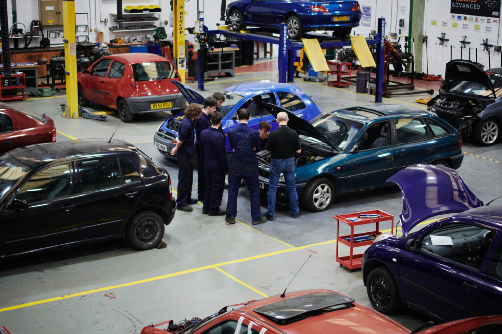 A group of male students in correct uniform watching a male tutor demonstrating working on a car engine in a garage environment at Walford College