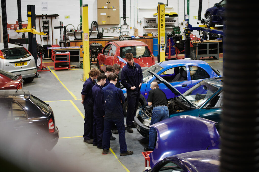 A group of male students in correct uniform watching a male tutor demonstrating working on a car engine in a garage environment at Walford College