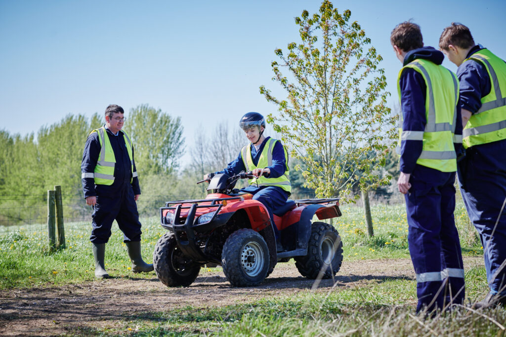 A male student in correct uniform and protection on a quad bike in the outdoors with a supervisor and other students nearby watching at Walford College